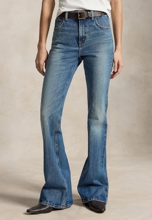 Femme portant un jean flare bleu taille haute avec poches avant, une ceinture noire, un haut blanc et des chaussures noires pointues, debout sur un fond neutre.
