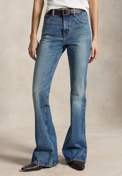 Mujer con vaqueros acampanados de talle alto y bolsillos delanteros, cinturón negro, top blanco y zapatos negros puntiagudos, de pie frente a un fondo neutro.