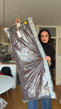 Dlouhý tmavě fialový oděv v plastovém obalu s visačkou velikosti "34". Má hladkou texturu a zahrnuje ramínko pro zavěšení.