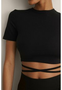Žena v přiléhavém černém crop topu s krátkými rukávy a křížovými popruhy obtočenými kolem pasu na jednobarevném pozadí.