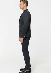 Egy sötétszürke, testhezálló öltönyt és fekete mokaszint viselő férfi profilból áll sima fehér háttér előtt.