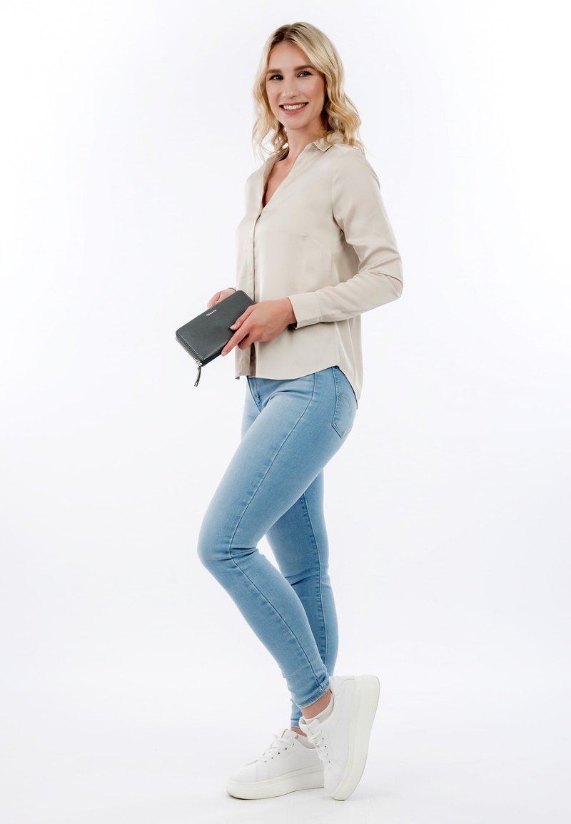 Smiling woman in beige blouse, light blue jeans, and white sneakers holding a dark gray wallet, standing against a white background.