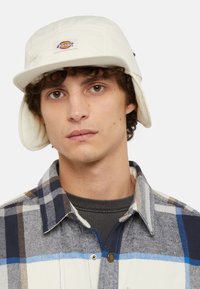 Weißer Bucket-Hut mit Ohrklappen, ausgestattet mit einer gesteppten Textur und einem runden Logo-Patch. Das Model trägt ein kariertes Hemd in Blau, Braun und Creme.