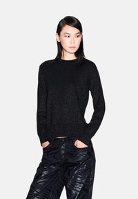 Zwarte trui met een glinsterende textuur, ronde halslijn en lange mouwen. Gecombineerd met glanzende, gemusterde zwarte broeken.