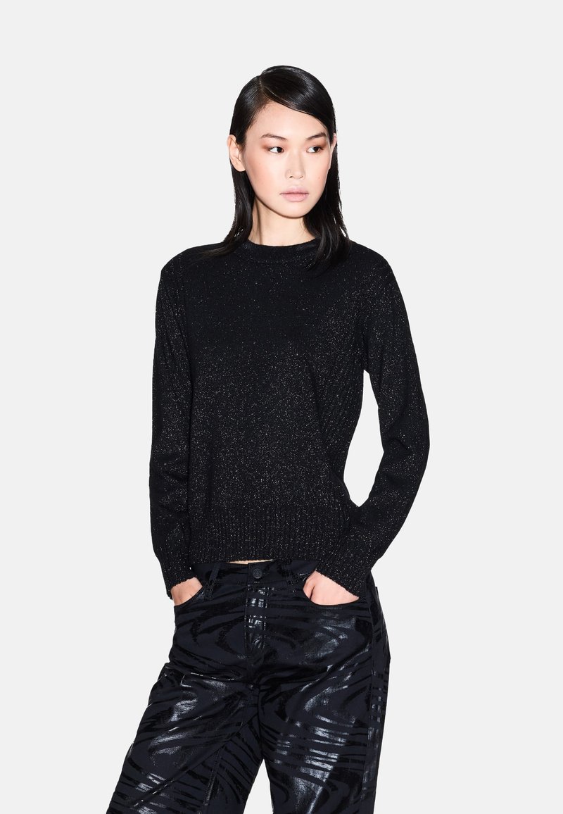 Zwarte trui met een glinsterende textuur, ronde halslijn en lange mouwen. Gecombineerd met glanzende, gemusterde zwarte broeken.