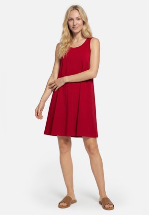 Frau mit langen blonden Haaren, die ein ärmelloses rotes knielanges Kleid und braune offene flache Sandalen trägt und vor einem weißen Hintergrund steht.