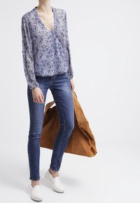 Chemisier à manches longues violet et blanc à motifs, associé à un jean skinny bleu et des chaussures blanches à enfiler, portant un grand sac en daim beige.