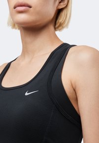 Κοντινό πλάνο του άνω κορμού με μαύρο αθλητικό τοπ Nike με ορατό λογότυπο, ανοιχτή επιδερμίδα, ξανθά κοντά μαλλιά, ουδέτερο φόντο.