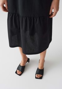 Schwarzes Kleid mit gerafftem Saum, geschichtetem Design und glatter Textur. Kombiniert mit schwarzen Leder-Mules mit offenem Zehenbereich und quadratischem Absatz.