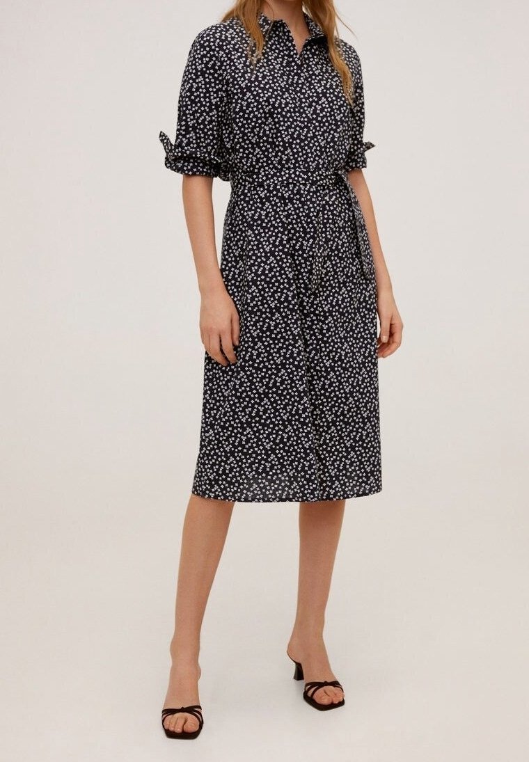 Femme portant une robe midi noire à imprimé floral blanc, manches retroussées et sandales à talons ouvertes noires, debout sur un fond uni.