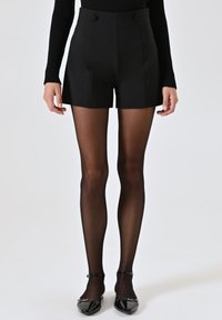 Hochtaillierte schwarze Shorts aus dehnbarem Stoff, mit zwei vorderen Knöpfen, kombiniert mit transparenten schwarzen Strumpfhosen und glänzenden schwarzen Schuhen.