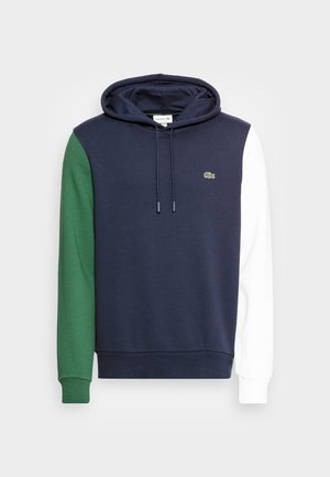 Hoodie bleu marine avec manche gauche verte, manche droite blanche, cordons frontaux et petit logo de crocodile vert sur la poitrine.