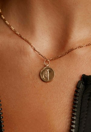 Collar de cadena de oro con un colgante redondo que presenta un diseño de figura en relieve, llevado sobre piel bronceada, junto a una tela negra con dientes de cremallera.