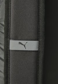 Schwarzer Textil-Rucksack mit glatter Oberfläche, ausgestattet mit einem reflektierenden grauen Streifen und kleinem Puma-Logo in der Mitte. Robuste Nähte sind sichtbar.
