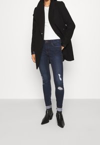 Abrigo negro sobre una blusa blanca de canalé, combinado con jeans ajustados rotos de un azul oscuro y botas negras de tobillo con punta.