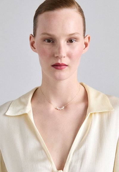 Χρυσή necklace με πέντε μικρές λευκές πέρλες, φορεμένη πάνω από μια ανοιχτοκίτρινη μπλούζα με υφή και γιακά.