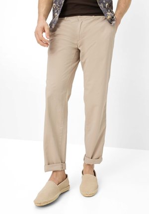 Mand iført beige opkørte bukser, beige slip-on sko, mørk skjorte og blå mønstret åben skjorte, stående mod hvid baggrund.