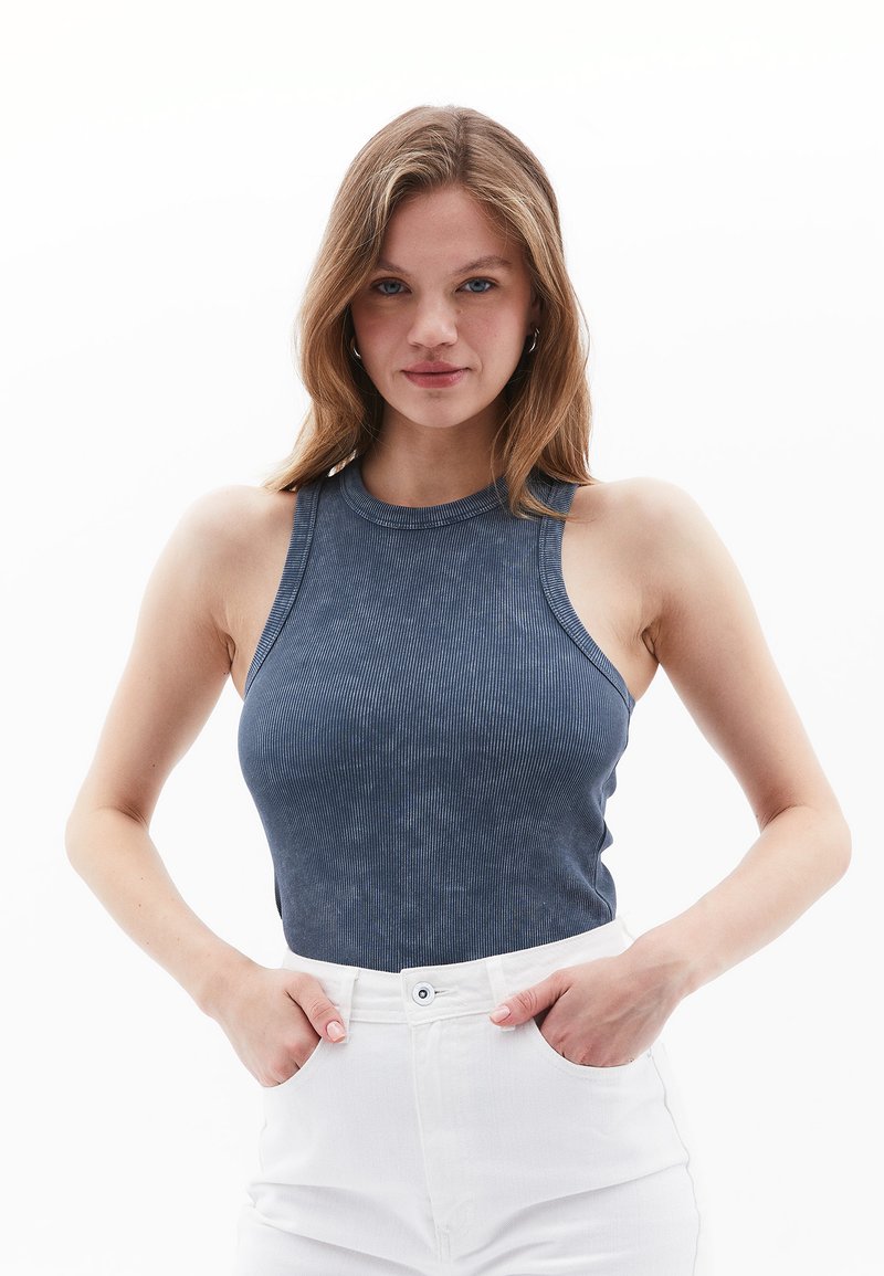Mørkeblå, ribbet ærmeløs top med høj hals og tætsiddende design. Parret med hvide højtaljede jeans, hænderne i lommerne.