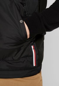 Czarna kurtka puchowa z prążkowanymi mankietami, wyposażona w boczną kieszeń oraz metkę z czerwono-biało-niebieskimi paskami na dole.