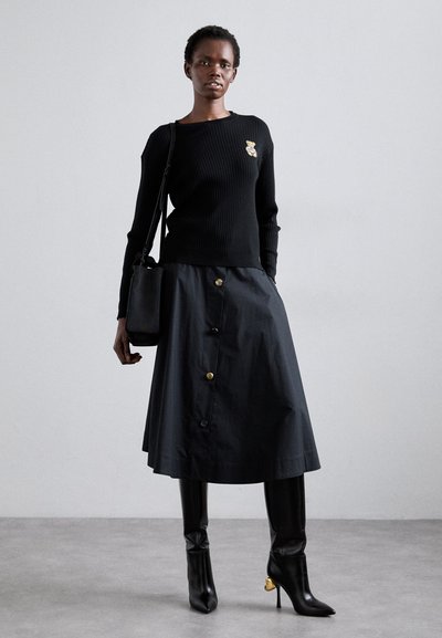 Pull noir côtelé avec une broche en forme d'ours, associé à une jupe noire mi-longue avec des boutons dorés et un petit sac à main noir, ainsi qu'à des bottes hautes.