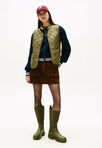 Frau trägt eine rote Mütze, eine olivgrüne gesteppte Weste, einen marineblauen Pullover, einen braunen Cordrock mit Taschen, schwarze Strumpfhosen und olivgrüne kniehohe Stiefel.