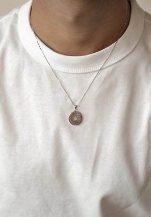Silberner Anhänger mit kreisförmigem Design und filigranen Mustern, der an einer dünnen Kette hängt und über einem weißen, gerippten T-Shirt getragen wird.