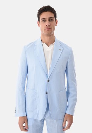 Uomo che indossa un blazer azzurro chiaro e pantaloni coordinati, sopra una maglia a V bianca, in piedi contro uno sfondo chiaro e uniforme.