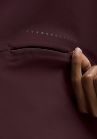 Burgundatygt med en blixtlåsficka, prytt av en rad små reflexpunkter ovanför. Hand som greppar blixtlåsets drag.