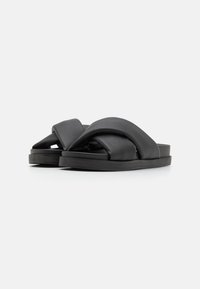 Sandales à glissière en cuir noir avec un design de sangle tordue, semelle plate et texture lisse. Esthétique minimaliste sans matériel ou accents visibles.