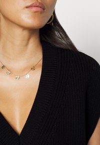 Schwarzes, geripptes Strickoberteil mit tiefem V-Ausschnitt, kombiniert mit einer filigranen Goldkette, die fünf kleine münzartige Anhänger enthält.