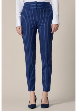 Kobieta ubrana w dopasowane niebieskie spodnie w prążki i niebieskie szpilki, z białą koszulą z długim rękawem wpuszczoną do środka.