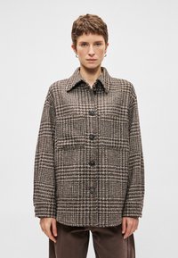 Karo jaku ar apkakli, brūnā un pelēkā krāsā, ar melniem pogām un brīvu piegriezumu, izgatavota no teksturēta auduma.