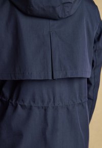 Vista posteriore di una giacca blu navy con cappuccio, con spalla ventilata e dettaglio in vita arricciato sul tessuto.