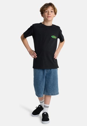 Junger Junge steht mit den Händen in der Hüfte, trägt ein schwarzes T-Shirt mit grünem Logo, Jeansshorts, weiße gestreifte Socken und schwarze Turnschuhe.