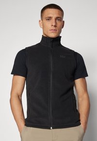 Gilet en polaire noir avec un col haut, fermeture éclair sur le devant et poches latérales. Porté par-dessus une chemise noire à manches courtes, texture lisse.