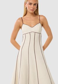 Rochie crem cu detalii maro, având un corset ajustat, decolteu în V, bretele subțiri și fustă evazată. Material neted cu cusături verticale.
