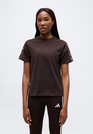 Vrouw draagt een donkerbruin Adidas T-shirt met luipaardprint strepen op de mouwen en bijpassende legging met wit Adidas-logo en zijstrepen.