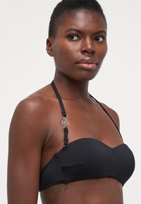 Crni bikini top s glatkim, prilagođenim dizajnom, podesivim trakama i metalnim akcentom na ramenu.