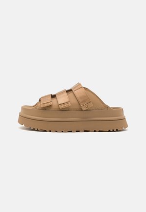 Infradito beige con tre cinturini regolabili, suola in gomma spessa e finitura texturizzata. Presenta un logo del brand ben visibile sul lato.