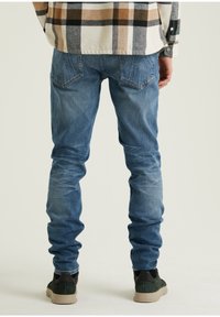 Modré džíny z denimu s úzkým střihem, s vybledlými oblastmi a zadními kapsami. Nošeny s tmavě zelenými teniskami, stojící na neutrálním pozadí.