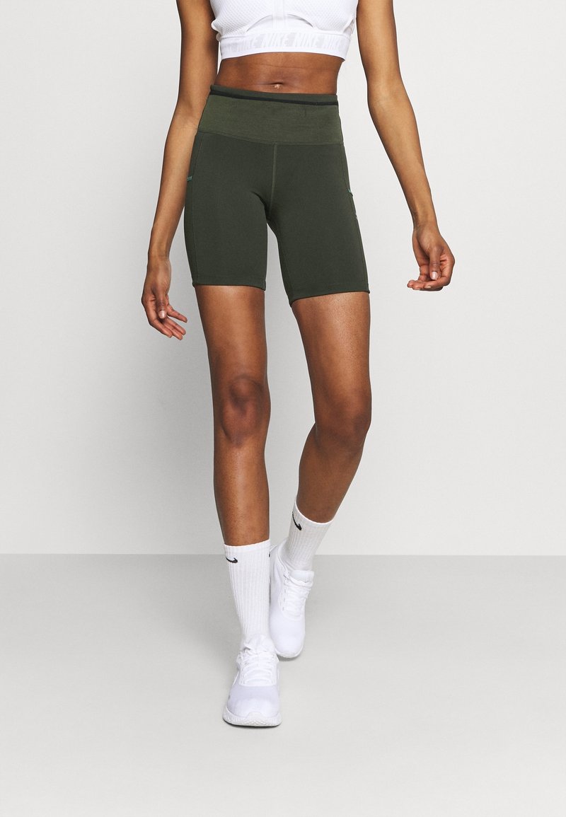Vrouw in donkergroene hoge fietsshorts, witte crop top, witte Nike-sokken en witte sportschoenen staat op een grijze vloer.