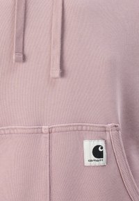 Lys rosa hettegenser med kengurulomme, snorer og et hvitt firkantet merke med en svart logo. Myk tekstur, uformelt design.