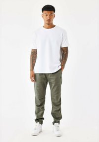 Camiseta blanca de manga corta con logotipo en relieve, combinada con pantalones tapered verde oliva con bolsillos y zapatillas blancas.