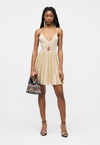 Vrouw met een mouwloze, V-hals beige en goud gestreepte jurk, die een met patronen versierde handtas vasthoudt en opvallende sandalen met hoge hakken draagt.