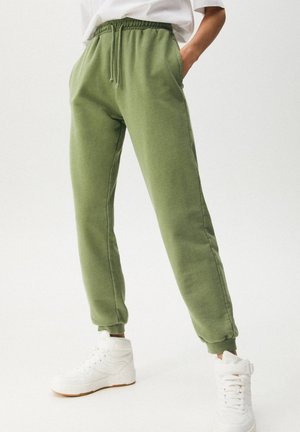 Person iført olivengrønne joggebukser med elastisk talje og manchetter, kombineret med hvide høje sneakers på en ensfarvet baggrund.