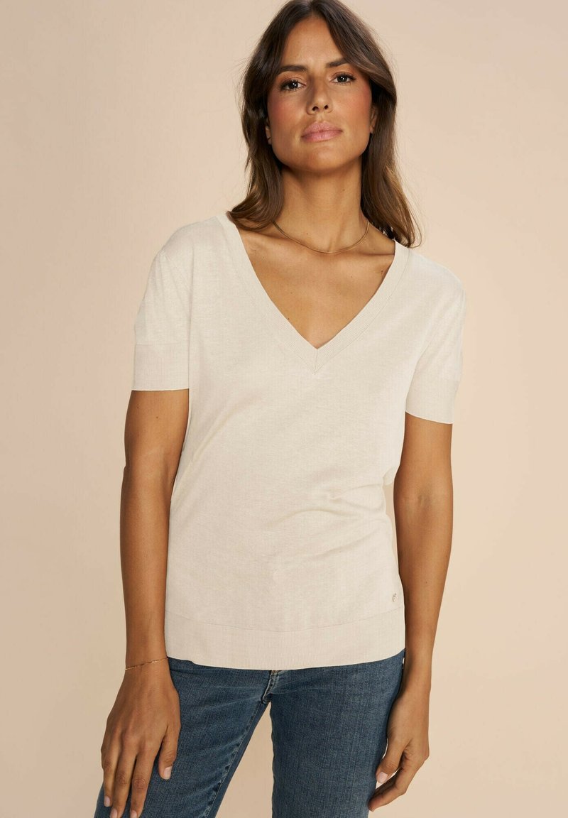 Ljust beige v-ringad topp med korta ärmar, tillverkad av mjukt tyg. Har en ribbad halsringning och fåll, ihop med mörkblå jeans.