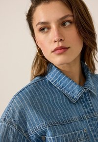 Chaqueta de mezclilla con rayas verticales, color azul claro, estilo abotonado, cuello puntiagudo y detalle de bolsillo en el pecho. Tejido suave y texturizado.