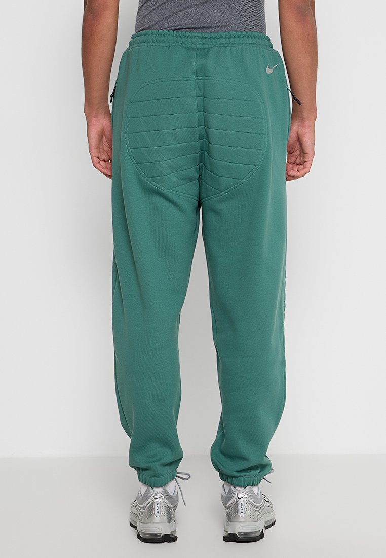 Vista posteriore di una persona che indossa pantaloni sportivi imbottiti verdi con vita e polsini elasticizzati, una maglietta grigia e scarpe da ginnastica argento, in piedi su un pavimento bianco.