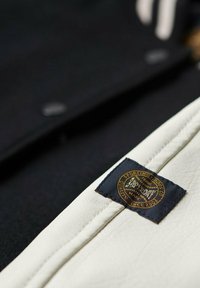 Svart jacka med vit läderdetalj, prydd med en dekorativ guld- och svart etikett samt metalliska detaljer. Slät textur.