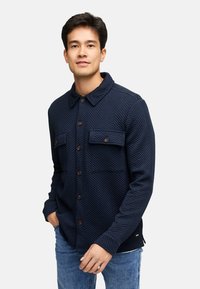 Navyblaue gesteppte Hemdjacke mit Fischgrätmuster, mit Kragen, zwei Brusttaschen und braunen Knöpfen. Getragen mit Jeans.